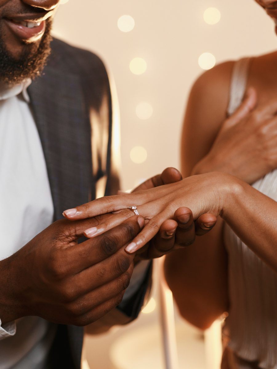 A Black man sliding an engagement ring onto his fiancee's finger, a Broom List engagement